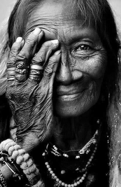 Beautiful the face of an older Indigenous woman wearing many necklaces and bracelets with one hand covering half her face. 